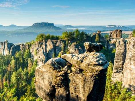 Blick von der Bastei in der Sächsischen Schweiz mit dramatischen Sandsteinsäulen über grünem Wald und fernen Tafelbergen unter blauem Himmel.