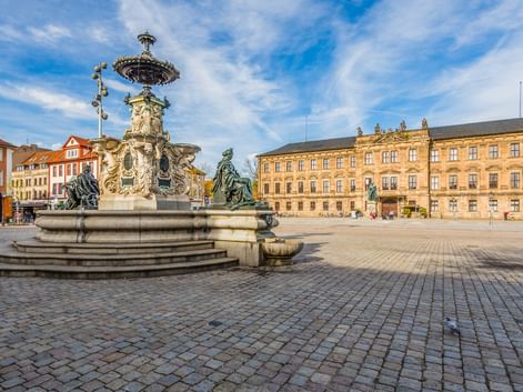 Prunkvoller Barockbrunnen mit Bronzestatuen auf dem Erlanger Schlossplatz. Großes gelbes Schlossgebäude im Hintergrund, Kopfsteinpflaster unter blauem Himmel.