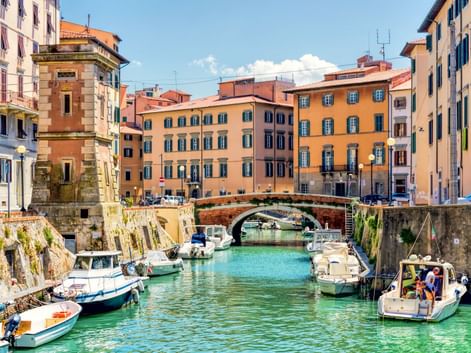Türkisfarbener Kanal in Venezia Nuova, Livorno mit festgemachten Booten entlang Steinmauern. Bunte Gebäude säumen beide Seiten, mit Steinbrücke in der Mitte.
