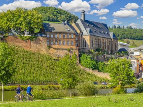 Zwei Radfahrer auf einem Weg neben einem Fluss mit der St. Laurentius Kirche auf einem Hügel in Saarburg, umgeben von Weinbergen und grüner Landschaft.