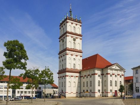 Weiße Stadtkirche mit hohem Turm und rotem Dach am Marktplatz in Neustrelitz, umgeben von historischen Gebäuden und Bäumen unter blauem Himmel.