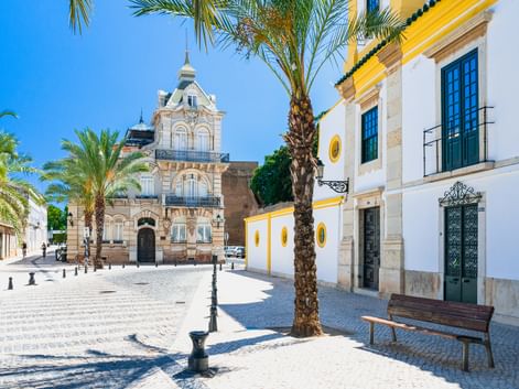Sonniger Kopfsteinpflasterplatz in Faro mit Palmen, historischer Kirche mit verzierter Fassade und weißen Gebäuden mit gelben Akzenten.