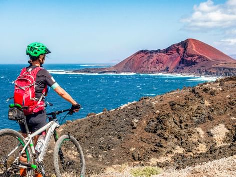Mountain biker with green helmet and red backpack on rocky terrain overlooking blue ocean and red volcanic cone in Lanzarote.