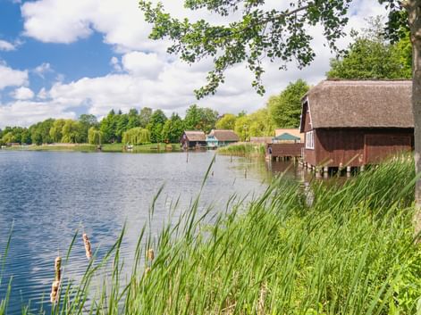 Traditionelles reetgedecktes Bootshaus auf Stelzen an einem ruhigen See mit Schilf im Vordergrund. Blauer Himmel mit Wolken und grüne Bäume säumen das Ufer.