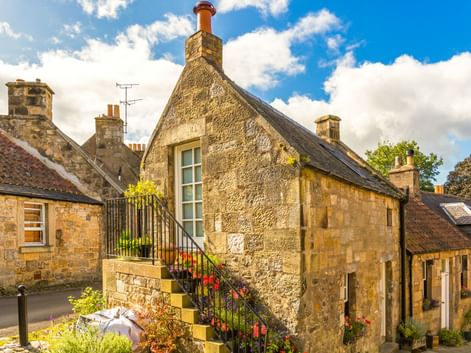 Traditionelle schottische Steinhäuser mit steilen Dächern und Schornsteinen in Falklands Altstadt. Bunte Blumen säumen die Steintreppe unter blauem Himmel.