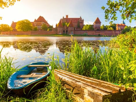 Marienburg in Malbork, Polen, über einen ruhigen Fluss mit blauem Boot und Holzbank im Vordergrund, eingerahmt von grünem Laub.
