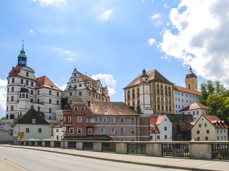 Blick auf die Neuburger Schlossanlage mit weißem Renaissanceschloss, Kirchturm und bunten historischen Gebäuden an der Donau unter blauem Himmel.