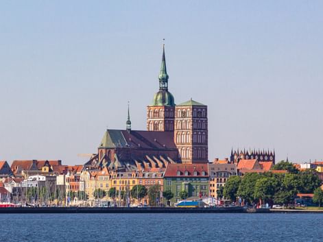 Blick über den Hafen von Stralsund mit bunten historischen Gebäuden am Ufer und der markanten St. Nikolaikirche mit ihrem Kupferturm.