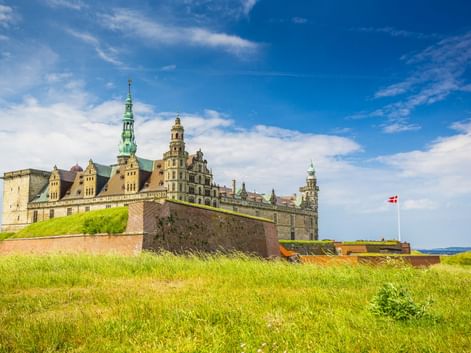 Schloss Kronborg in Helsingør mit grünen Kupfertürmen und Renaissance-Türmen, umgeben von Grashängen. Dänische Flagge weht an hohem Mast.