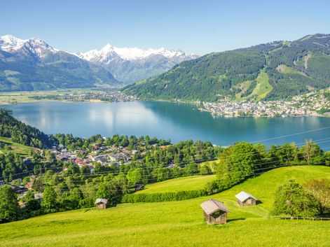 Luftaufnahme des Zeller Sees umgeben von grünen Wiesen mit Holzhütten, bewaldeten Bergen und schneebedeckten Alpengipfeln unter blauem Himmel.