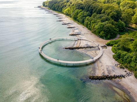 Luftaufnahme eines kreisförmigen Holzstegs, der ins Meer in Aarhus ragt. Die Struktur bildet einen vollständigen Kreis mit Besuchern.