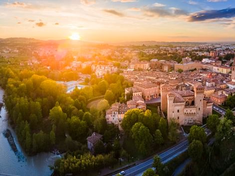 Luftaufnahme der Burg Rocca di Vignola umgeben von Bäumen und historischer Stadt bei Sonnenuntergang. Links fließt ein Fluss durch grüne Landschaft.