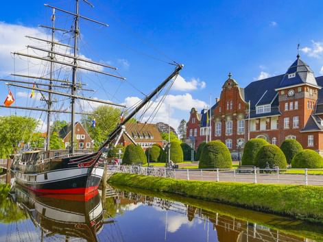 Historisches Segelschiff im Kanal vor dem roten Backsteinrathaus. Runde Hecken säumen das Ufer unter blauem Himmel in Papenburg.