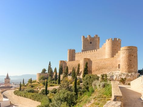 Burg La Atalaya in Villena mit Steinmauern und Türmen auf einem Hügel, umgeben von Zypressen und einem gewundenen Weg unter klarem blauem Himmel.