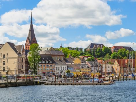 Blick auf den Flensburger Hafen mit bunten historischen Gebäuden, Kirchturm und Booten am Ufer unter blauem Himmel mit weißen Wolken.