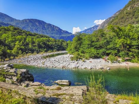 Klarer Fluss mit felsigen Ufern im Maggiatal, umgeben von grünen Bäumen und steilen Bergen unter blauem Himmel. Kleiner Strand mit Menschen sichtbar.