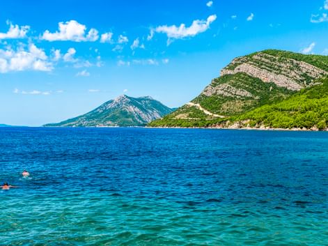 Zwei Schwimmer im klaren türkisfarbenen Wasser nahe der Halbinsel Pelješac. Grüne bewaldete Berge erheben sich von der Küste unter blauem Himmel.