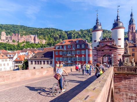 Cyclist on Heidelberg's Old Bridge with twin gate towers, historic buildings, and Heidelberg Castle on forested hillside in background.