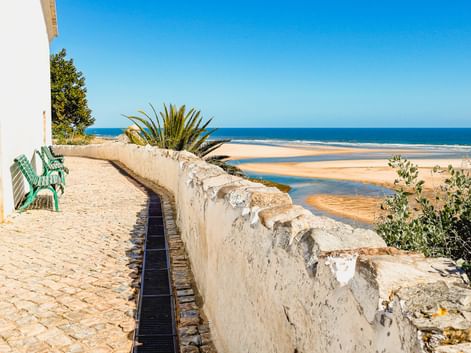 Kopfsteinpflaster-Promenade mit grüner Bank und weißer Mauer in Cacela Velha mit Blick auf Sandstrand und blaues Meer unter klarem Himmel.