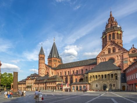 Mainzer Dom mit roten Sandsteintürmen am Domplatz unter blauem Himmel. Historische Gebäude und Fußgänger auf dem Platz.