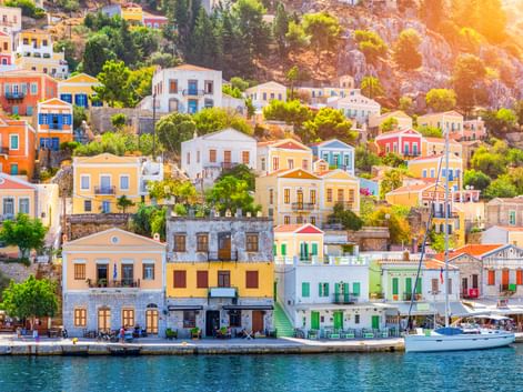 Bunte neoklassizistische Häuser in Gelb, Weiß, Blau und Orange säumen die Uferpromenade der Insel Symi mit einem Boot im Hafen.