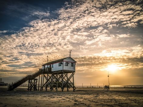 Hölzernes Pfahlhaus mit Treppe am Nordseestrand bei Sonnenuntergang. Dramatischer bewölkter Himmel mit goldenem Sonnenlicht.