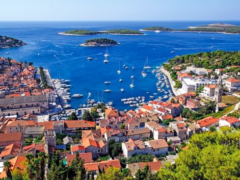 Luftaufnahme des Hafens von Hvar in Kroatien mit rotgedeckten Gebäuden, zahlreichen Booten im blauen Wasser und grünen Inseln in der Ferne.