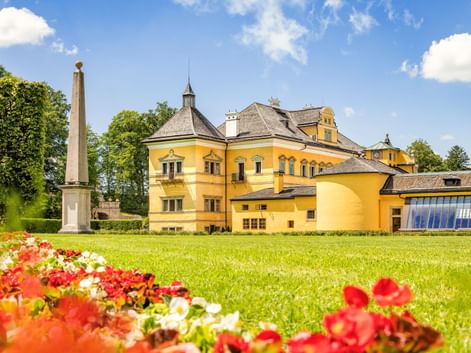 Gelbes Schloss Hellbrunn in Salzburg mit gepflegten Rasenflächen, bunten Blumenbeeten und einem Obelisken unter blauem Himmel mit weißen Wolken.
