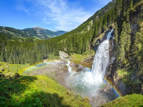 Krimmler Wasserfälle stürzen über Felsklippen mit Regenbogen im Sprühnebel, umgeben von grünem Alpental und bewaldeten Bergen in Österreich.