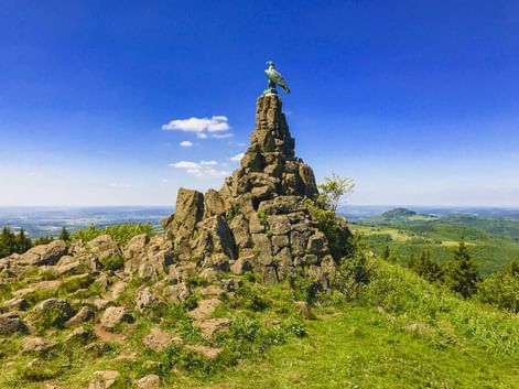 Steinpyramide mit Adlerskulptur auf dem Gipfel der Wasserkuppe, mit Blick auf grüne Hügel unter blauem Himmel.