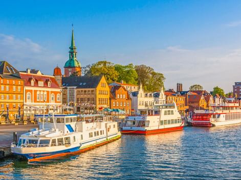 Kappeln Hafen mit weißen und roten Booten an bunten Ufergebäuden. Eine Kirche mit grünem Turm erhebt sich hinter orangen und gelben Häusern.