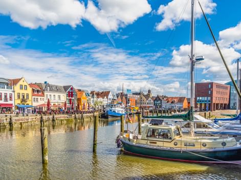 Husumer Hafen mit bunten historischen Häusern entlang der Uferpromenade. Segelboote und Yachten liegen im Hafen unter blauem Himmel mit weißen Wolken.