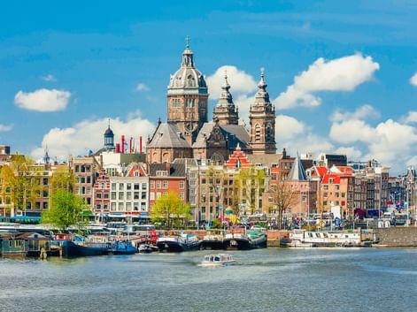 Amsterdamer Waterfront mit den Türmen der St. Nicolaaskerk über bunten Grachtenhäusern. Hausboote säumen den Hafen unter blauem Himmel.