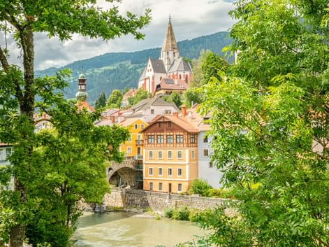 View of Murau old town in Austria with a prominent church spire, colorful historic buildings along a river, framed by green trees and mountains.