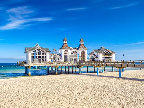 Historisches weißes Seebrückengebäude mit verzierten Türmen in der Ostsee bei Sellin. Sandstrand im Vordergrund unter blauem Himmel.