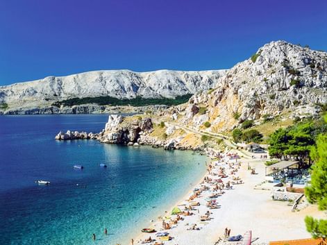 Kieselstrand mit Badegästen in türkisfarbener Bucht auf Insel Krk bei Baska. Weiße Felsengebirge erheben sich hinter dem klaren Adriawasser.