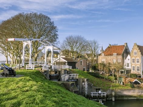 Weiße hölzerne Zugbrücke mit traditionellen niederländischen Häusern entlang eines Kanals in Schoonhoven. Grüne Grasuferböschung und kahle Bäume unter blauem Himmel.