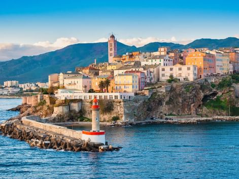 Rot-weißer Leuchtturm auf Steinmole im Hafen von Bastia mit bunten Altstadthäusern auf Klippe und Bergen im Hintergrund.
