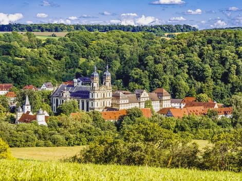 Panoramablick auf Kloster Schöntal mit Doppeltürmen, umgeben von rotgedeckten Gebäuden, eingebettet in grüne bewaldete Hügel in Hohenlohe.