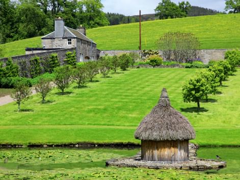 Strohgedeckter Pavillon auf Teich im Hercules Garden bei Blair Castle. Grüne Rasenfläche mit Bäumen führt zu Steingebäude, Hügel dahinter.