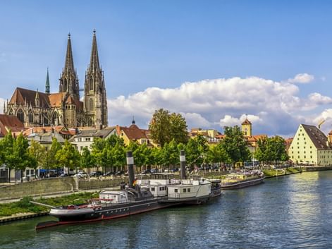 Regensburg Cathedral with twin Gothic spires overlooking the Danube River. Historic boats moored along the waterfront with colorful buildings and trees.