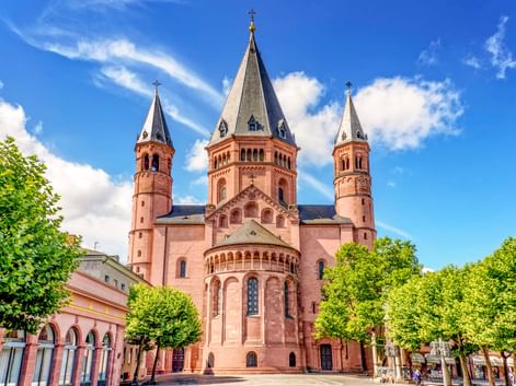 Mainz Cathedral with three red sandstone towers topped with crosses, flanked by green trees under a blue sky with white clouds.