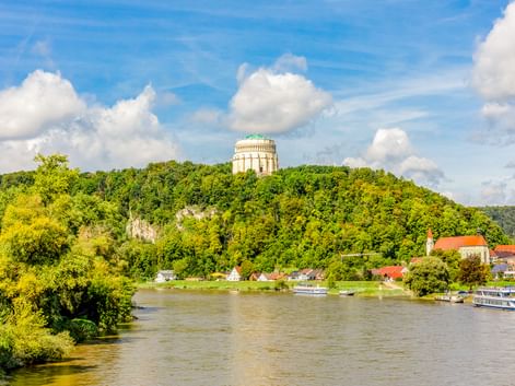 The Befreiungshalle monument sits atop a forested hill above Kelheim. The Danube River flows below with boats moored along the green riverbank.