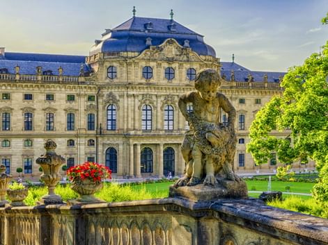 Würzburger Residenz mit prunkvoller Barockfassade und blauem Dach. Steinbrunnen mit Skulptur im Vordergrund, umgeben von grünem Rasen und Bäumen.