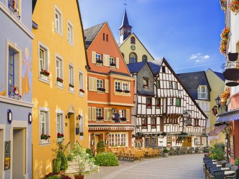 Bunte Fachwerkhäuser und bemalte Fassaden umgeben einen sonnigen Marktplatz in Bernkastel-Kues mit Außengastronomie und Kirchturm.