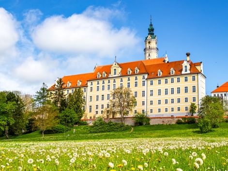 Benedictine Abbey in Donauwörth with yellow facade and orange roof, surrounded by green meadow with yellow dandelions and white flowers.