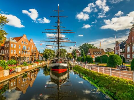 Museumssegelschiff mit hohen Masten liegt in einem Kanal in Papenburg, flankiert von traditionellen Backsteingebäuden und gepflegten Gärten unter blauem Himmel.