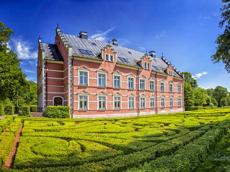 Palsjö Schloss, ein rosa Barockschloss mit grauem Dach, umgeben von ornamentalem Heckenlabyrinth unter blauem Himmel bei Helsingborg.