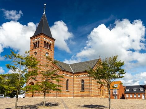 Backsteinkirche Erlöserkirche in Esbjerg mit hohem Spitzturm und Rundbogenfenstern, umgeben von jungen Bäumen auf einem gepflasterten Platz.