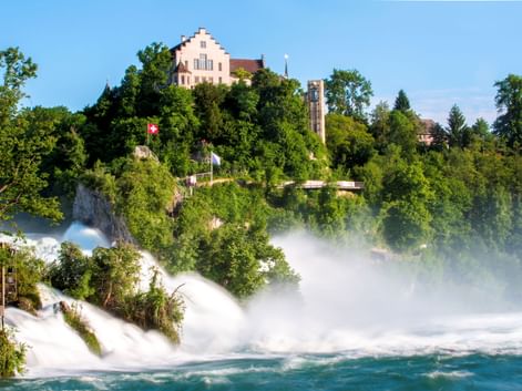 Rheinfall in Schaffhausen mit kraftvoll strömendem Wasser, Gischt und Regenbogen. Schloss Laufen thront auf grünem Hügel mit Schweizer Flagge.